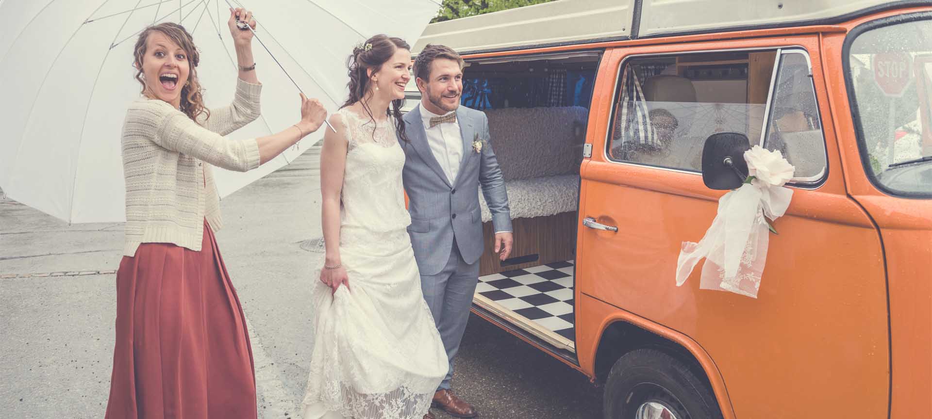 lachendes Hochzeitspaar steigt in alten VW-Bus mit Chauffeur und schwarz-weiss karriertem Boden ein