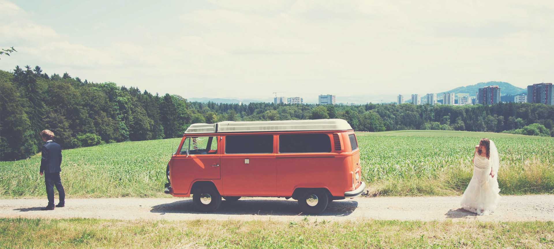 Braut versteckt sich als Überraschung hinter orangem Oldtimer-Büssli vor Berner Skyline