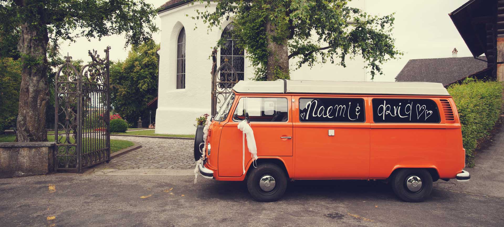 geschmücktes Oldtimer VW-Bus Hochzeitsfahrzeug mit Chauffeur wartet vor Kirche auf Brautpaar