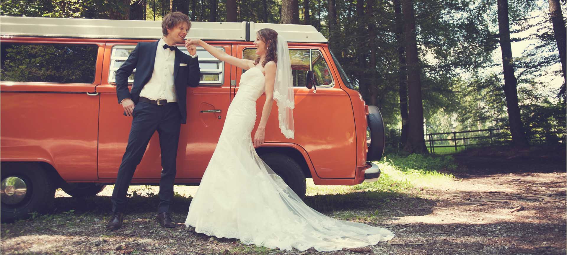Le marié embrasse main de la mariée souriante devant combi VW vintage dans forêt bernoise